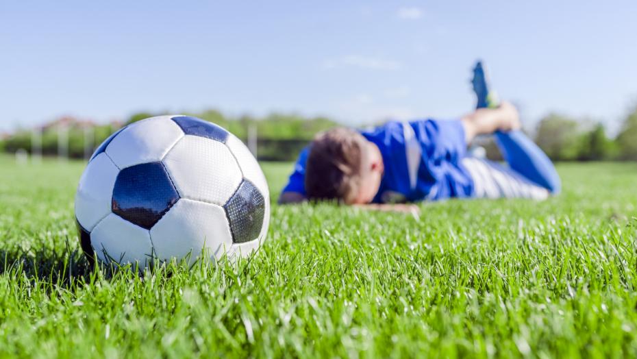 Man lying on stomach in field and holding ankle with soccer ball in foreground