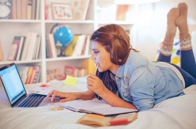 Teenager with pencil in mouth lying on bed with laptop open nearby